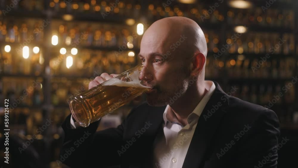 A man dressed in a white shirt and a jacket is drinking beer at a pub. The manager is relaxing at the bar after work. Close-up shot in profile. A man dressed in a white shirt and a jacket is drinking beer at a pub. The manager is relaxing at the bar after work. Close-up shot in profile.