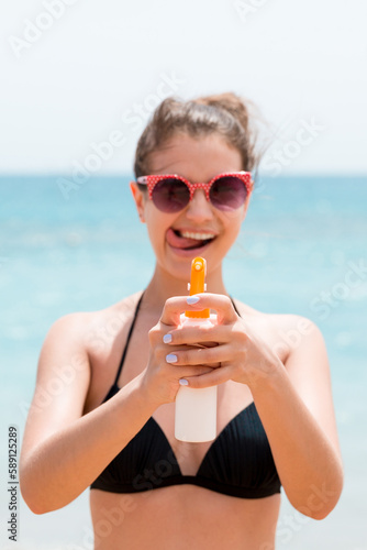 Young woman is holding a spray of sun cream for body at the camera at the beach
