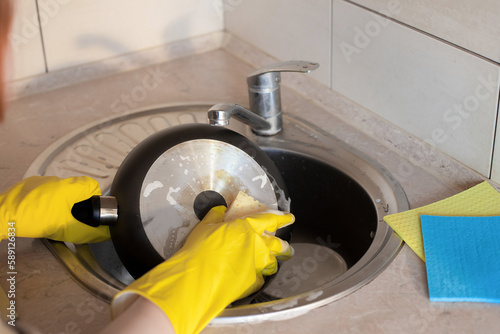 Women's hands, in yellow rubber gloves, wash a large dirty frying pan with a sponge and soap under running water under a tap. The plot is about housework and cleanliness in the kitchen.