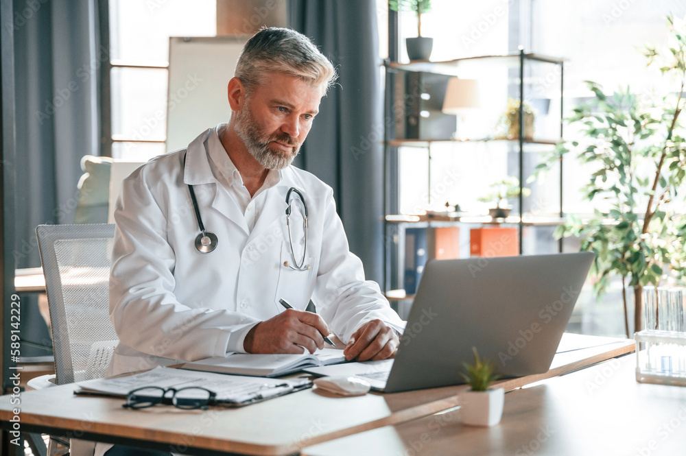 Sitting by table with laptop, writing data into document. Senior doctor in white coat is at work indoors Sitting by table with laptop, writing data into document. Senior doctor in white coat is at work indoors