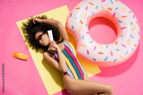top view of african american model in swimwear and trendy sunglasses holding sunscreen near inflatable ring on pink.