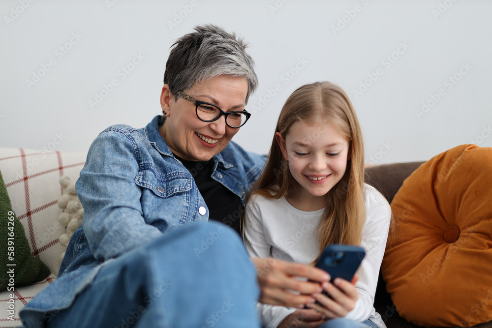 Happy grandmother and granddaughter watching fun content on phone in home interior. Happy grandmother and granddaughter watching fun content on phone in home interior.