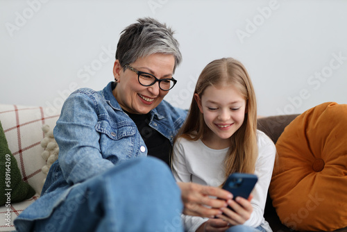 Happy grandmother and granddaughter watching fun content on phone in home interior.