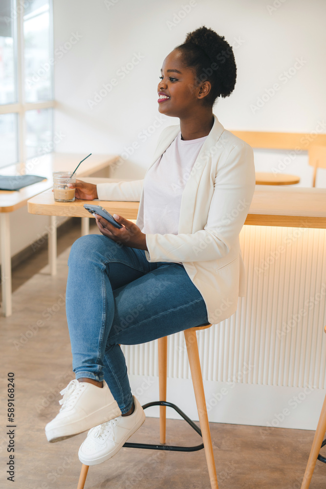 Cheerful beautiful african woman using an app in her smartphone device holding ice coffee sitting ina city cafe. Internet information. Delicious breakfast Cheerful beautiful african woman using an app in her smartphone device holding ice coffee sitting ina city cafe. Internet information. Delicious breakfast