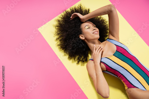 top view of satisfied african american model in colorful striped swimsuit lying on yellow and pink background.