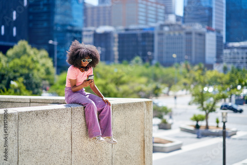 Young woman using smart phone on ledge in sunny city