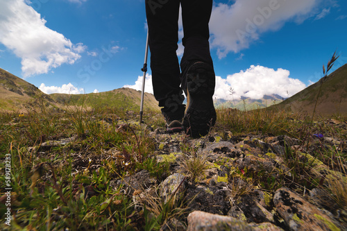 Hiking trail with flowers, green grass and stones. Close up of hiking boots in the mountains against the backdrop of mountains and fluffy clouds