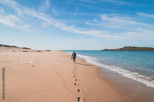 Passionate backpacker, backpack and boots in hand, walks along the Praia da Ilha do Pessegueiro beach on the Atlantic Ocean near Porto Covo, Portugal. In the footsteps of Rota Vicentina