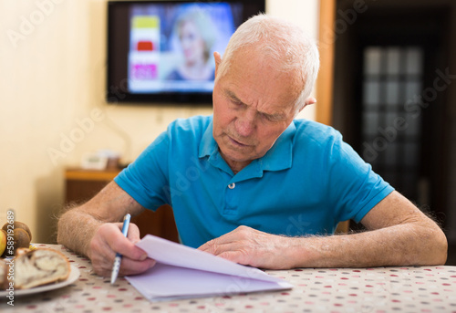 Serious older man writing letter at table in living room