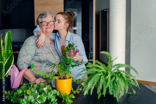 Loving teen granddaughter and grandmother kissing and embracing, replanting house plants in a pots in kozy kitchen at home. Special moments together. an attractive woman visiting granny. © Iryna