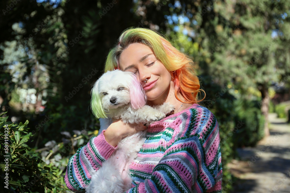Young woman wearing colorful knitted sweater at the park hugging her maltese dog. Female with multicolored hair and her pup with color matched ears. Copy space, background. Young woman wearing colorful knitted sweater at the park hugging her maltese dog. Female with multicolored hair and her pup with color matched ears. Copy space, background.