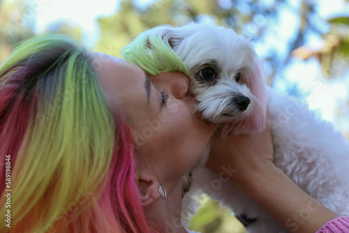 Close up shot of a young woman with multicolored hair kissing her beloved maltese dog. Female and her pup with color matched ears. Copy space, background.