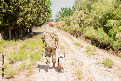 Unrecognizable, slender male fisherman in camo outfit walking down dirt road with obedient spotted dog in summer. Back view of adult guy with fishing pole going along bushy path. Concept of hobby.