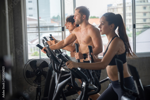 Group of young shirtless man and woman in sportswear working on elliptical machine for cardio training in modern gym against cityscape while looking outside window during morning for healthy lifestyle