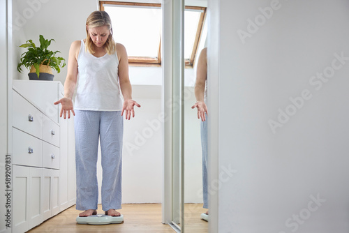 Menopausal Mature Woman Concerned With Weight Gain Standing On Scales In Bedroom At Home photo