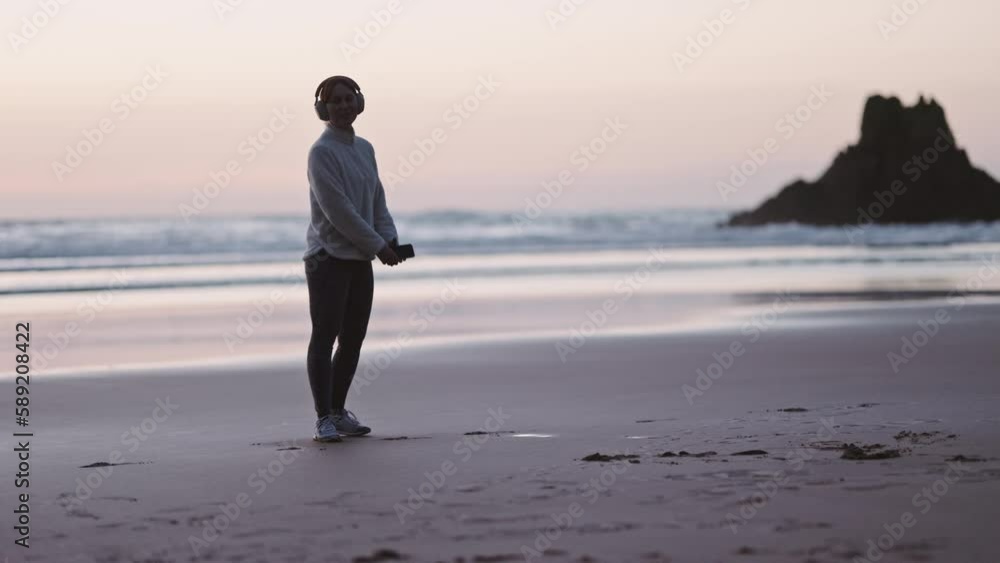Woman in headphones dancing at the ocean beach at sunrise. Happy Woman with smartphone listens to music after a workout enjoying herself near the sea. Cinematic freedom healing Woman in headphones dancing at the ocean beach at sunrise. Happy Woman with smartphone listens to music after a workout enjoying herself near the sea. Cinematic freedom healing