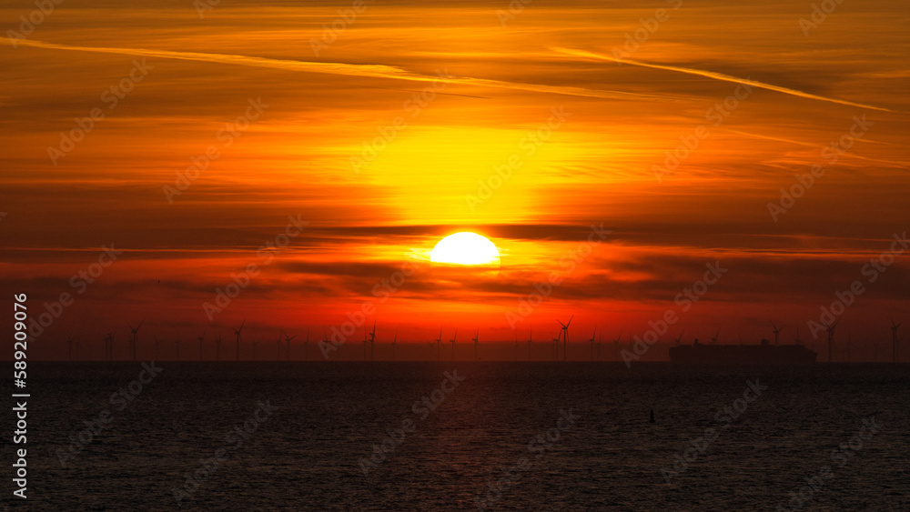 Die Sonne versinkt als glühender Ball wie flüssiges Gold dramatisch in der Nordsee mit Silhouetten von Windkraft Anlagen am Horizont