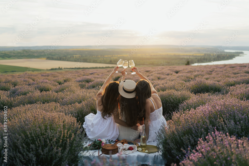 Back view of three female friends in nature clinking with a glasses of wine. Picnic in lavender field at sunset.