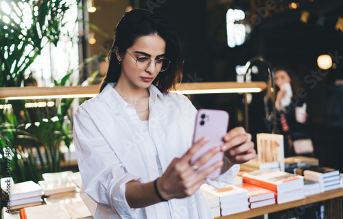 Young woman standing and taking selfie in bookshop with smartphone
