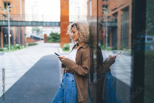 Black female with cellphone leaning on glass wall outdoors