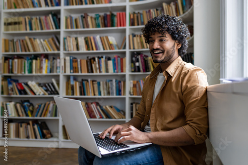 Young hispanic student studying inside academic library among bookshelves, man typing on laptop keyboard, smiling contentedly sitting near window.