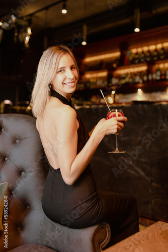 Portrait of young well-groomed slim woman wearing black dress with open back with fair hair sitting on arm of brown sofa in restaurant, holding glass with red cocktail with straw, posing. Vertical.