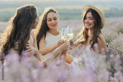 Obraz na płótnie A beautiful happy young girlfriends clinking glasses with white wine at summer picnic in a lavender field. w salonie