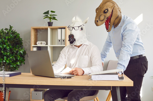Two funny business men wearing animal masks standing at their workplace with laptop and having discussion in office, considering new projects or startups, analyzing company. Fools day concept.