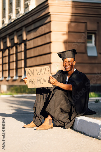 Happy black guy sitting with cardboard poster on street looking for job. University or college graduating student in graduate gown and cap. Study and future career, uncertainty, unemployment problem.