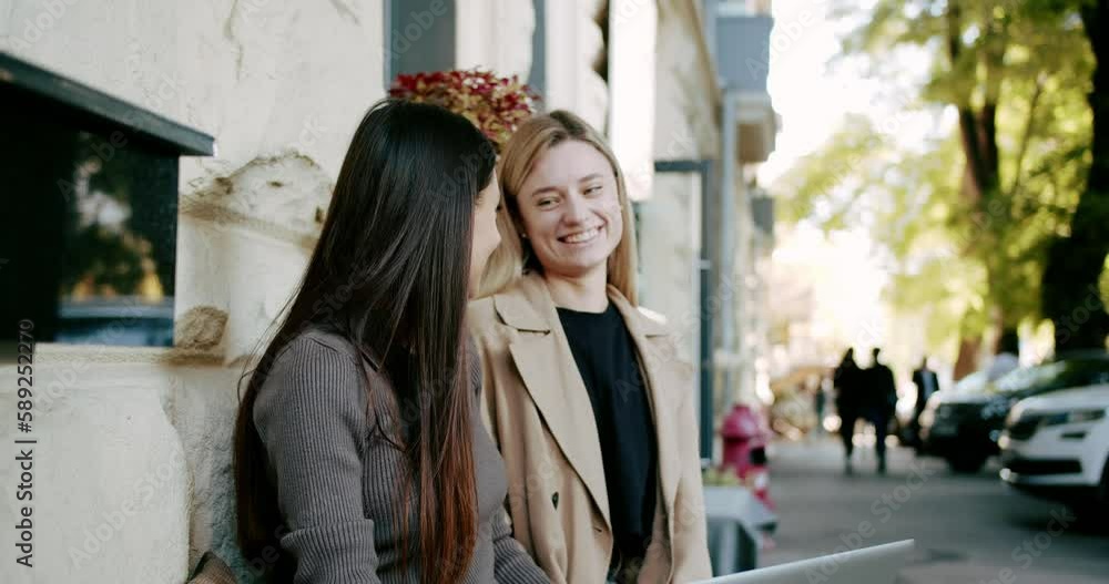 Young women with coffee chatting on street. Handheld shot of young girlfriends with coffee to go talking and laughing while spending sunny weekend day on city street together