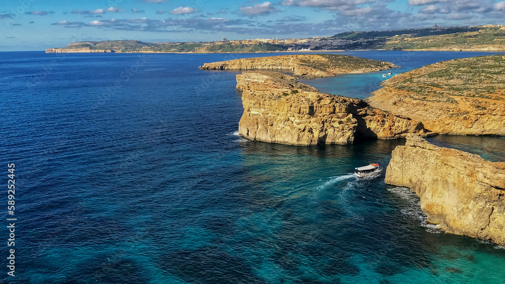 Small boat in the background strolling through the blue waters of blue lagoon in malta, with the rocky landscape around.