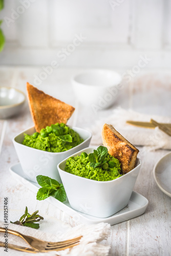 Two bowls of pea puree with basil, mint and roasted bread on white wooden background, vertical, copy space