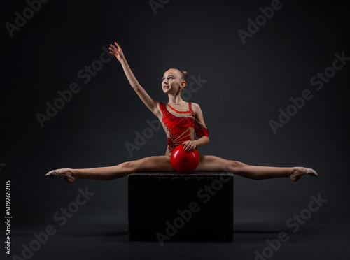 Young rhythmic gymnast girl posing in red leotard with red ball in dark studio