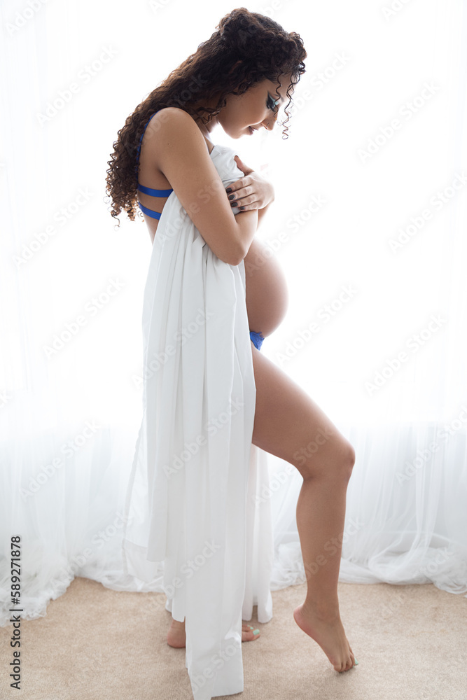 Lifestyle portrait of a mixed race black women standing in front of a window and wrapped in a white sheet. She is pregnant and is looking down at her pregnant belly. This was taken in a bedroom. Lifestyle portrait of a mixed race black women standing in front of a window and wrapped in a white sheet. She is pregnant and is looking down at her pregnant belly. This was taken in a bedroom.