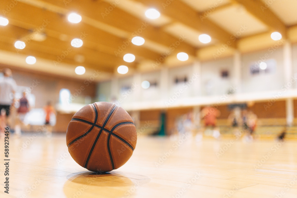 Naklejka premium Classic Orange Basketball Ball On Wooden Sports Court. Junior Level Basketball Players Playing Game in Blurred Background. Basketball Training Background.