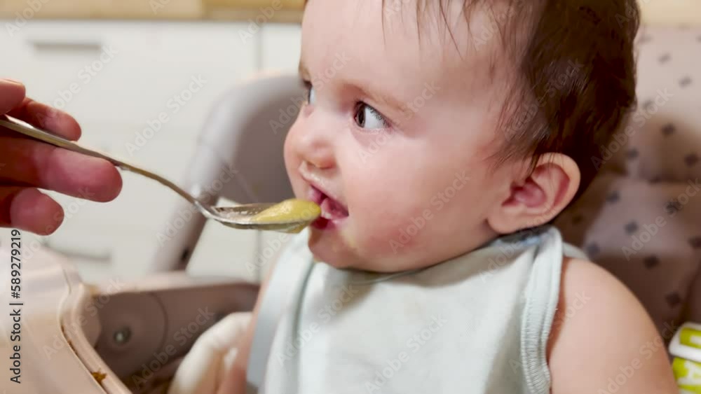 mom feeds baby at the table. dirty a baby newborn dirty mouth eats from a spoon. happy family kindergarten proper nutrition newborn. baby eats fruit puree lifestyle from a spoon mom feeds baby at the table. dirty a baby newborn dirty mouth eats from a spoon. happy family kindergarten proper nutrition newborn. baby eats fruit puree lifestyle from a spoon