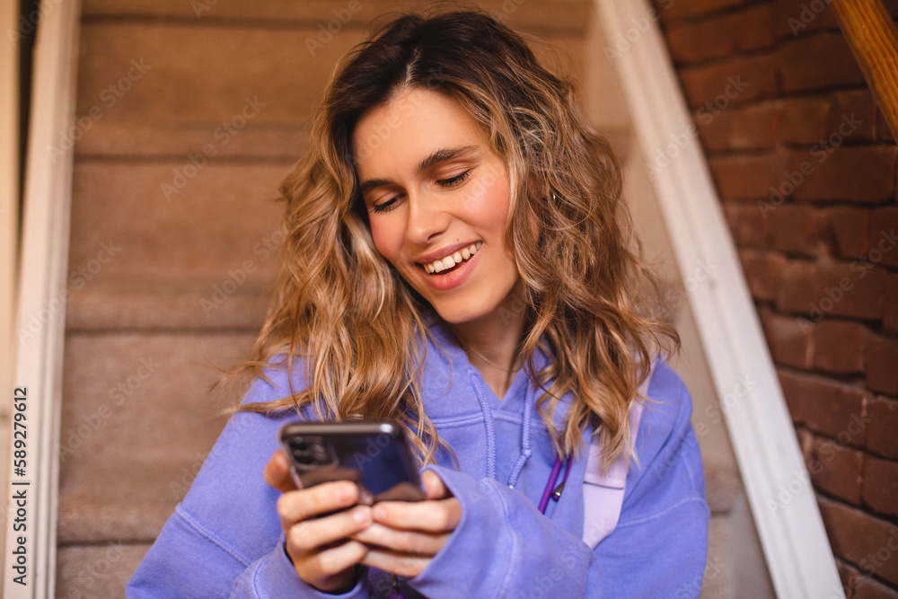 Young woman at home in near stairs in a warm purple hoodie, happy hold mobile, smiling, cozy evening. Lifestyle, female beauty concept. Blonde curly woman in house read messages and look happy. Young woman at home in near stairs in a warm purple hoodie, happy hold mobile, smiling, cozy evening. Lifestyle, female beauty concept. Blonde curly woman in house read messages and look happy.