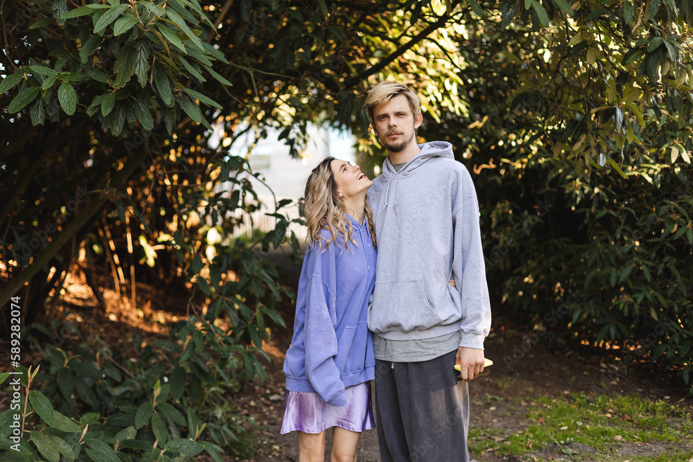 Fototapeta premium Couple walk outdoors in the park. Woman warped man and look at him laughing. Girl wear purple hoody and skirt, man wear grey hoody and pants. Fashion outfit of couple, love relationship.