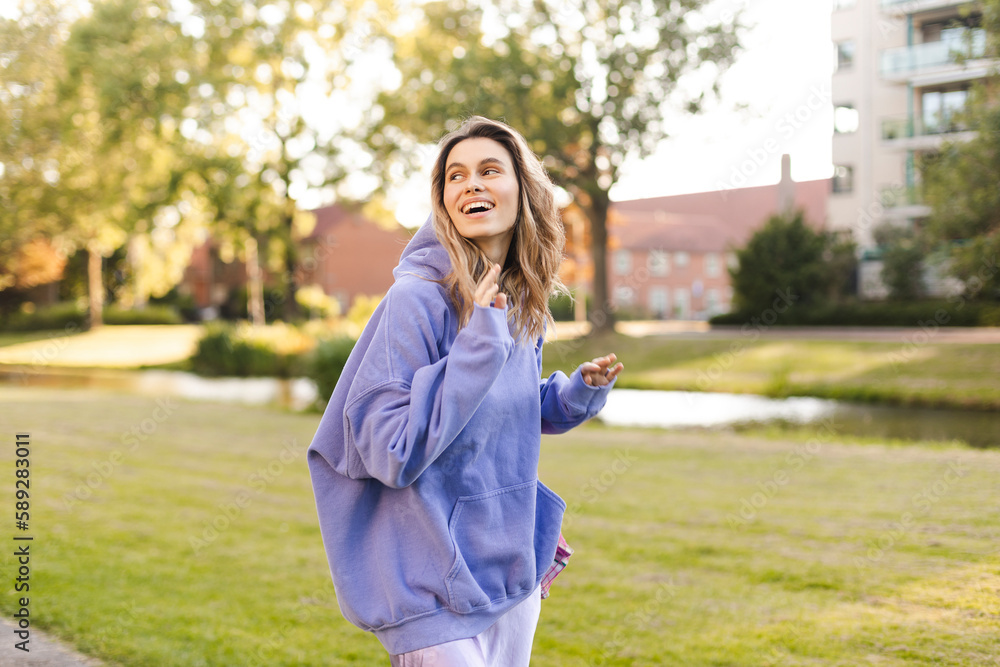 Pretty young caucasian woman with curly blonde hair turns around walking in the park. Blonde smiling, dressed in casual purple hoody and skirt. Rest and recovery concept. Happy girl hold pink tote bag
