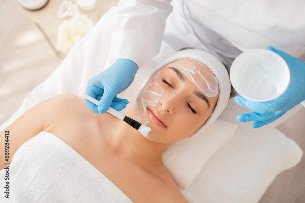Naklejka premium Skincare Treatment. Cosmetologist Applying Moisturising Mask On Face Of Beautiful Young Woman