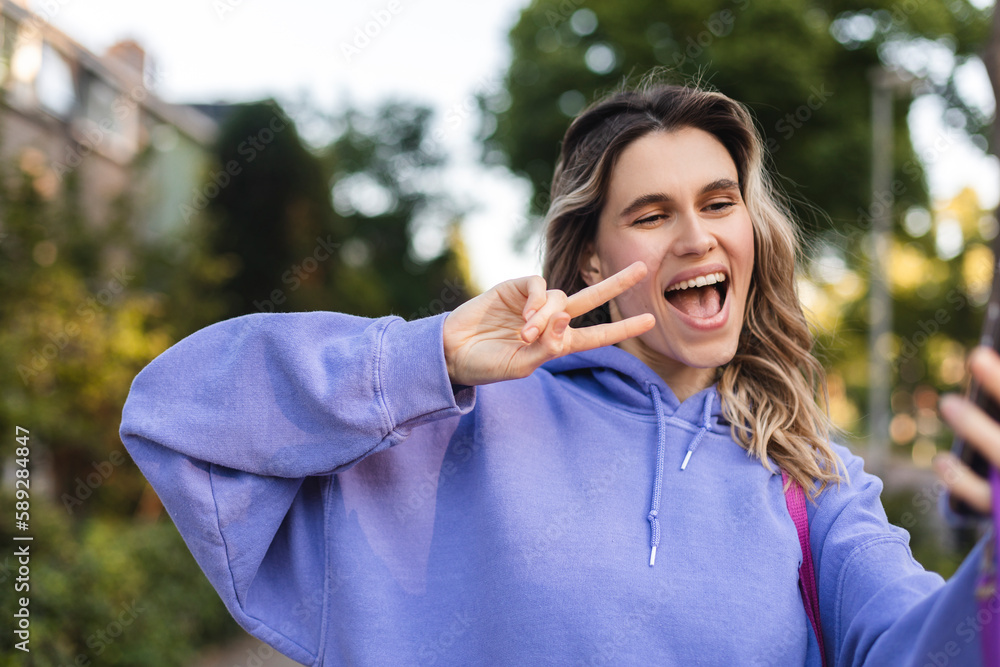 Happy young woman photographing herself using her mobile phone. Caucasian female taking selfie with her smart phone at city. Positive lady making photo outside in purple hoody and show peace v sign. Happy young woman photographing herself using her mobile phone. Caucasian female taking selfie with her smart phone at city. Positive lady making photo outside in purple hoody and show peace v sign.