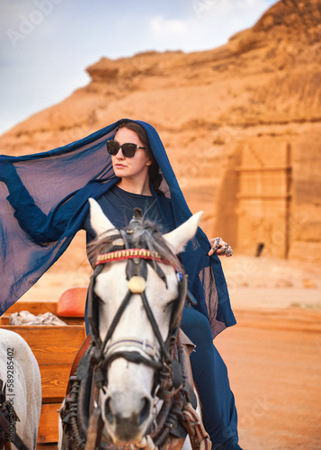 Young woman with dark long dress, head scarf and sunglasses, sitting on a horse, blurred Hegra or Mada’in Salih ruins in background - female tourist travel influencer like photo from Saudi Arabia
