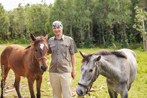 Fototapeta Naklejka Na Ścianę i Meble -  Smiling, young man standing next to brown horse foal, another animal near, blurred forest background