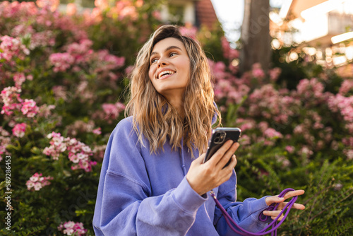 Attractive blonde curly woman wear purple hoody hold mobile phone and look at side, look happy and smiling. Amazing enjoy woman walk near blossom flowers.