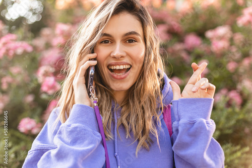 Wallpaper Mural Smiling modern woman wear purple hoody with crossed fingers speaking on a smartphone on bloom flowers background. Hopeful woman waiting for good news in a mobile phone call. Torontodigital.ca