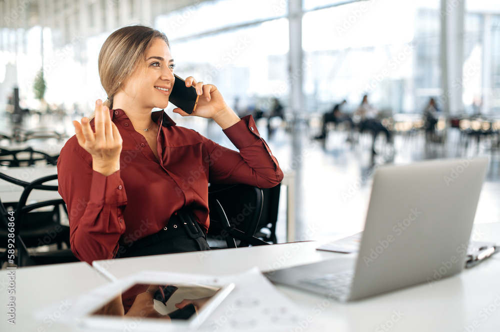 Positive beautiful elegant blonde caucasian woman in a formal shirt, office employee, manager, sit at her work desk in the office, talking on a smartphone with colleague or client, looking away, smile Positive beautiful elegant blonde caucasian woman in a formal shirt, office employee, manager, sit at her work desk in the office, talking on a smartphone with colleague or client, looking away, smile