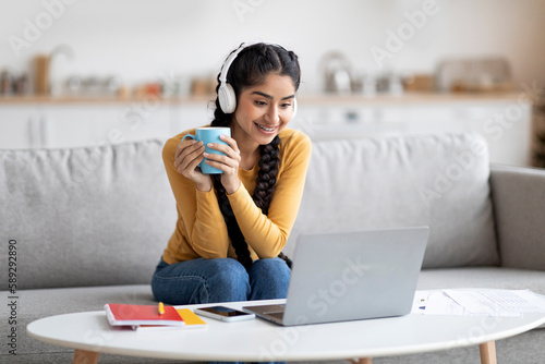Cheerful indian woman watching videos on laptop and drinking coffee at home