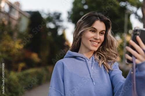 Single adult woman wear purple hoody uses her cell phone to stay connected, take photos, selfies, video calls, locate herself on the map to share experiences. Curly blonde woman walk on the street.