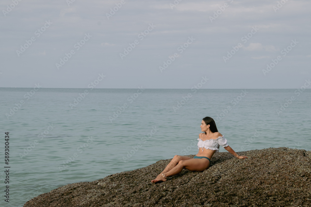 a young girl in a swimsuit is sitting on the sand on the seashore by the rocks