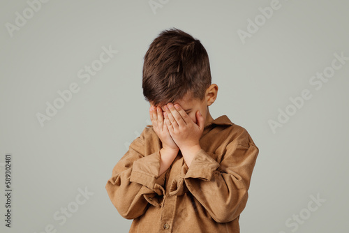 Sad caucasian 6 years old little kid covering face with hands, crying isolated on gray studio background photo